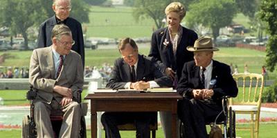 President George H.W. Bush siging the ADA Act of 1990 while seated at a table surrounded by four people.