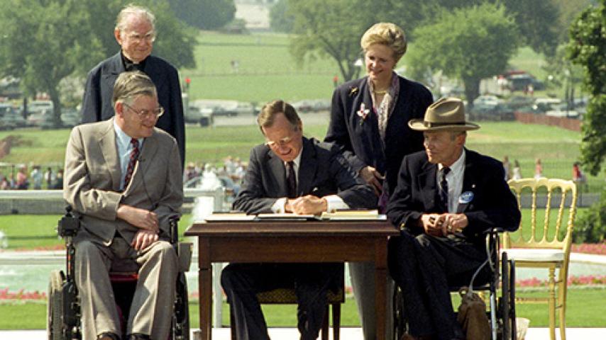 President George H.W. Bush siging the ADA Act of 1990 while seated at a table surrounded by four people.